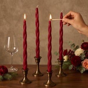May include: Four tall, deep red twisted taper candles standing in antique brass-toned metal candle holders. Two of the candles are lit with visible flames, and one is being lit by a match. A clear glass goblet and floral arrangement are nearby.