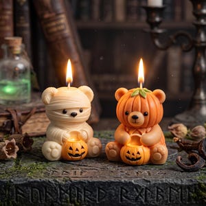 Puede incluir: Dos velas con forma de oso con mechas encendidas. La vela de la izquierda es un oso color crema envuelto en vendas como una momia, sosteniendo una pequeña calabaza naranja. La vela de la derecha es un oso marrón claro con un sombrero de calabaza.
