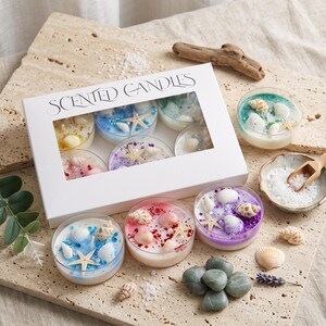 以下が含まれることがあります： SCENTED CANDLESと書かれた白い長方形の箱には、小さな貝殻、ヒトデ、カラフルなグリッターで飾られたワックス入りの丸いガラス容器が6個入っています。箱の外には、さらに3個のキャンドルとバラバラの貝殻が置かれています。