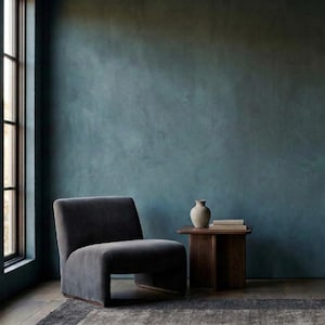 May include: A modern interior scene featuring a dark gray armchair, a small wooden side table with a vase and books, and a floor lamp. The room has a teal-colored wall and a patterned rug. A large window is on the left.