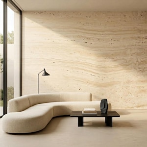 May include: A curved cream-colored sofa sits in a room with a large beige travertine stone wall. A black rectangular coffee table holds a dark abstract sculpture and two books. A black floor lamp stands behind the sofa near a glass wall.