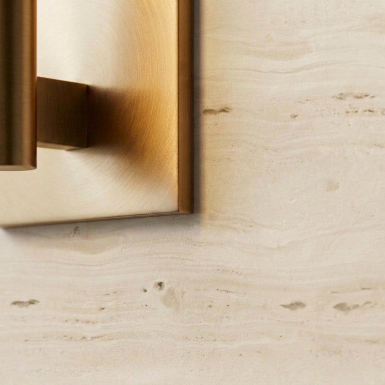 May include: Close-up of a brass wall sconce with a rectangular backplate and cylindrical arm. The sconce has a warm, metallic finish. The background is a light beige stone wall with subtle veining.