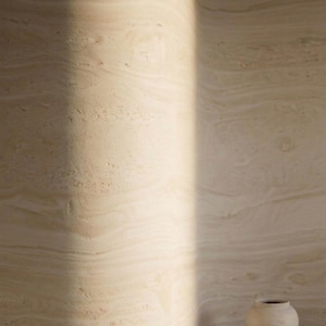 May include: A minimalist interior scene featuring a light-toned, curved wall with a natural, layered texture. A small, off-white ceramic vase sits on the floor. The scene is bathed in soft, natural light, creating a serene and artistic ambiance.