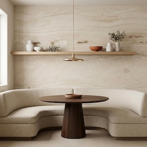 May include: A modern dining area featuring a round, dark wood table with a pedestal base, a curved beige sofa, and a light fixture. A wooden shelf displays decorative bowls and vases against a textured wall.