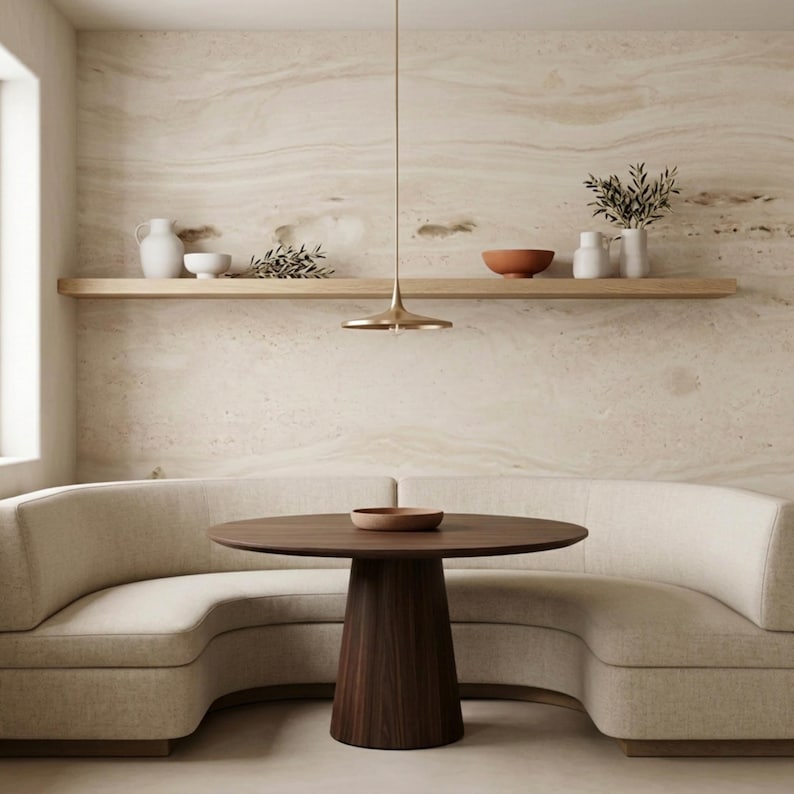 May include: A modern dining area featuring a round, dark wood table with a pedestal base, a curved beige sofa, and a light fixture. A wooden shelf displays decorative bowls and vases against a textured wall.