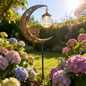 Puede incluir: Un estaca de jardín de metal con un marco de media luna con recortes de estrellas y una figura de colibrí. Una bombilla esférica de vidrio craquelado cuelga de la parte superior de la luna, emitiendo un brillo cálido sobre un poste fino.