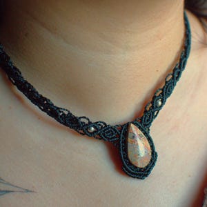 May include: A black macrame necklace featuring a central teardrop-shaped stone with mottled brown, tan, and cream colors. The cord is intricately knotted with small silver-toned beads integrated into the pattern on both sides of the pendant.