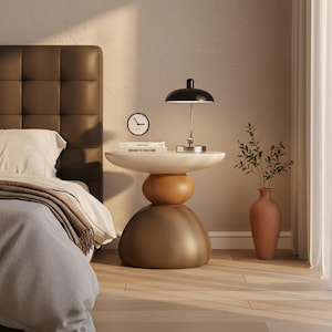May include: A sculptural side table featuring a stacked design with a white circular top, a rounded amber middle section, and a large dark brown dome-shaped base. The table holds a black dome-shaped lamp, a small round clock, and a stack of books.
