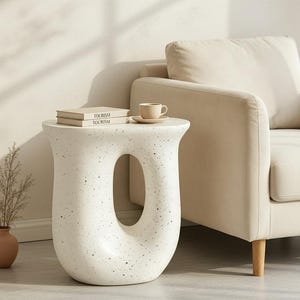 May include: A white side table with a speckled finish and a large central oval cutout. The table has a smooth, curved silhouette and a wide top surface. It is constructed from a solid material with a matte texture, featuring a distinct hollow center.