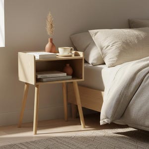 May include: A light wood nightstand with four tapered legs and an open storage shelf. The top surface holds a small terracotta vase with a dried plant, a ceramic cup on a saucer, and a book. The shelf contains two stacked books and a small vase.