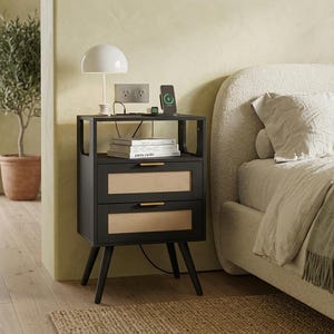 May include: A black nightstand with two drawers featuring light-colored woven cane panels and thin gold-toned horizontal handles. The nightstand has an open top shelf, four tapered black legs, and a flat surface for a lamp and charging devices.