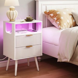May include: A white nightstand with two drawers featuring vertical ribbed detailing and gold-toned handles. The unit includes an open top shelf illuminated by integrated purple LED lighting, supported by four tapered legs, and connected to a power cord.