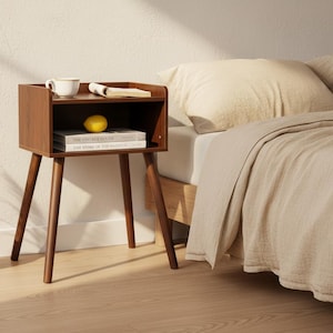 May include: A dark brown wooden nightstand with four tapered legs and an open storage shelf. The top surface holds a white ceramic mug and an open book. The shelf contains a yellow lemon resting on two stacked books titled The Vintage House.
