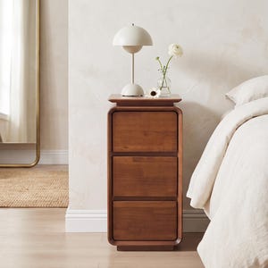 May include: A tall, rectangular wooden nightstand featuring three stacked drawers with rounded corners. The piece is constructed from a medium-toned wood with a visible grain pattern and sits on a base that matches the rounded frame design.