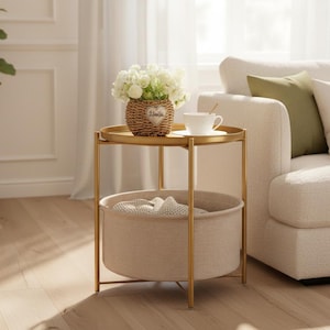 May include: A round gold metal side table with a tray top and a lower beige fabric storage basket. The table holds a woven basket with white flowers and a white ceramic teacup. The basket features a small heart-shaped tag with text.