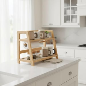 May include: A two-tiered light wood kitchen organiser featuring a paper towel holder on the middle level. The top shelf holds three ceramic mugs, while the bottom shelf contains three small glass jars and two additional ceramic mugs.