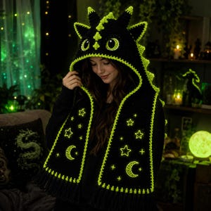May include: A black crochet dragon hood and scarf set with neon green detailing. The hood has dragon features, and the scarf is decorated with glowing stars and crescent moons. The set is finished with a fringe along the bottom edge.