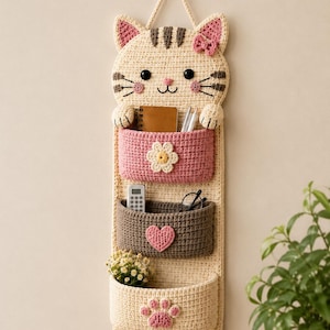 May include: A beige and pink crochet cat-shaped wall organizer with three pockets. The cat has a bow, whiskers, and a striped tail. The pockets are pink, brown, and beige, decorated with a flower, heart, and paw print. The organizer is hanging on a wall.