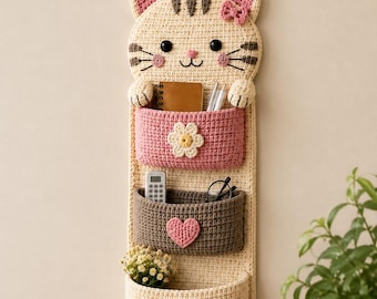 May include: A cream-colored crocheted wall organizer shaped like a cat with three pockets. The top pocket is pink with a white flower, the middle is brown with a pink heart, and the bottom is cream with a pink paw print. It features a striped tail.