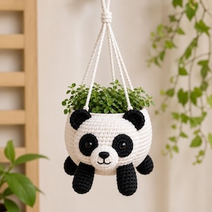 May include: A white and black crocheted panda hanging planter featuring a round body, two black ears, two black eye patches with dark eyes, a small snout with a stitched nose and mouth, and four black limbs, suspended by white braided ropes.