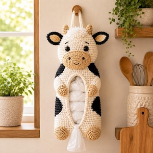 May include: A crochet cow-shaped plastic bag dispenser featuring a cream-coloured body with black spots, tan hooves, and tan horns. The dispenser has a central opening to store bags and a bottom opening for dispensing, hanging from a wooden wall hook.