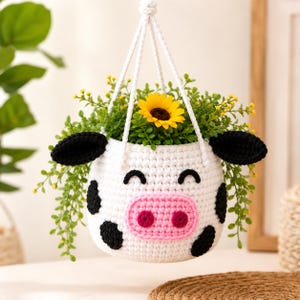 May include: A white crocheted hanging planter shaped like a cow with black spots, black ears, and a pink snout. The planter features a smiling face with closed eyes and contains a green plant with a single yellow sunflower bloom.