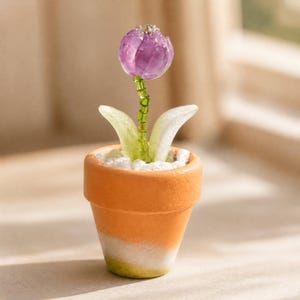 Pode incluir: Uma flor em vaso em miniatura com uma cabeça de tulipa de vidro roxo translúcido, um caule feito de pequenas contas verdes e duas folhas de vidro verde-pálido. A planta está num pequeno vaso de barro cor de terracota cheio de pedras brancas.