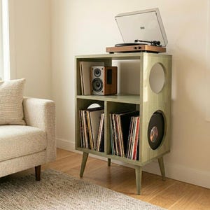 Puede incluir: Un mueble tocadiscos verde salvia con un tocadiscos en la parte superior, un altavoz y discos de vinilo. El mueble tiene un diseño moderno de mediados de siglo con recortes redondeados y patas anguladas. El mueble es de madera y tiene un acabado liso.