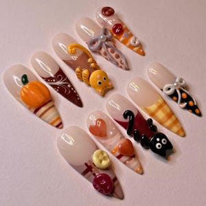 Puede incluir: Un juego de diez uñas postizas con diseños de temática otoñal que incluyen una calabaza naranja en 3D, un gato marrón, un gato negro, lazos, corazones y detalles circulares. Las uñas muestran patrones de rayas, cuadros y lunares en tonos naranja, marrón, negro y crema.