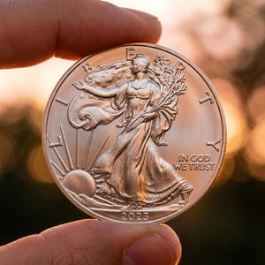 Może przedstawiać: Srebrna moneta trzymana między palcami, z reliefem kobiety w powiewającej szacie, trzymającej gałązkę. Moneta ma napis "IN GOD WE TRUST" i rok "2023". Tło jest rozmyte, z ciepłymi kolorami zachodu słońca.