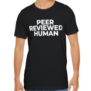 Può includere: T-shirt nero con la scritta bianca "PEER REVIEWED HUMAN" sul petto. La maglietta è a girocollo e sembra essere realizzata in un materiale morbido. La persona che indossa la maglietta è in piedi davanti a uno sfondo bianco.