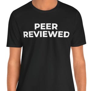 Può includere: T-shirt nera con la scritta "PEER REVIEWED" stampata in grassetto bianco maiuscolo. Il testo è centrato sul petto. La maglietta ha girocollo e maniche corte.