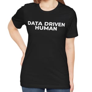 Puede incluir: Camiseta negra con el texto blanco "DATA DRIVEN HUMAN". La camiseta está hecha de un material suave y tiene cuello redondo. La modelo lleva vaqueros azules.