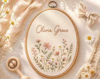 Puede incluir: Un bastidor de bordado ovalado de madera que contiene tela blanca con el nombre Olivia Grace bordado en hilo marrón, rodeado por un colorido patrón floral con flores rosas y blancas con tallos y hojas verdes.