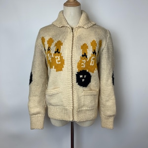 May include: A cream-colored, chunky knit zip-up cardigan featuring a bowling-themed design. The front displays yellow bowling pins with white numbers and a black bowling ball. The sleeves have black circular patches at the elbows.