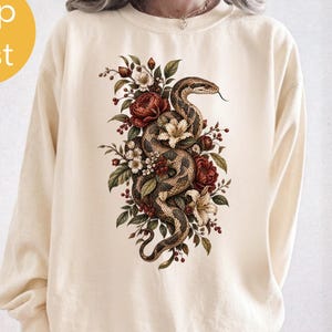May include: A cream-coloured crewneck sweatshirt featuring a central graphic print of a coiled snake intertwined with various flowers, including red roses and white lilies, surrounded by green leaves and small red berries on the front chest area.
