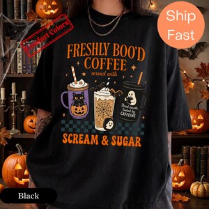 May include: A black t-shirt featuring a graphic print with the text FRESHLY BOO'D COFFEE served with SCREAM & SUGAR. The design includes three beverage containers: a purple mug with a black cat in a pumpkin, a glass with a spiderweb, and a black mug.