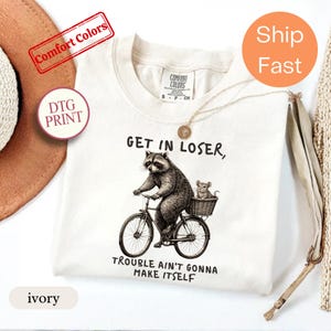 May include: An ivory t-shirt featuring a black ink illustration of a raccoon riding a bicycle with a mouse sitting in a rear basket. The text above reads "GET IN LOSER," and the text below reads "TROUBLE AIN'T GONNA MAKE ITSELF." The shirt has a Comfort Colors brand label.