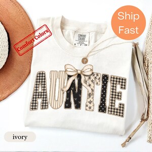 May include: An ivory short-sleeved t-shirt featuring the word AUNTIE in large block letters with varied black and beige patterns including gingham, stripes, and polka dots. A beige ribbon bow is centred over the letter N on the shirt.