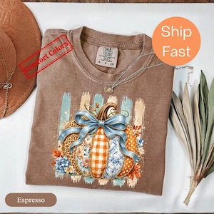 Pode incluir: Uma t-shirt cor de espresso com um gráfico de uma abóbora decorada com padrões azuis, laranja e brancos, incluindo estampas de leopardo, xadrez e florais. Um grande laço azul fica no topo da abóbora, rodeada por folhas de outono.
