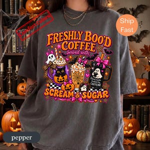 Könnte beinhalten: Ein anthrazitfarbenes T-Shirt mit einer bunten Grafik von drei Kaffeetassen im Halloween-Design. Der Text lautet FRESHLY BOO'D COFFEE served with SCREAM & SUGAR. Das Motiv zeigt eine schwarze Katze, Geister, Kürbisse, Fledermäuse und Spinnennetze.