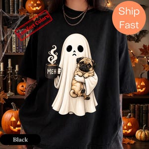 Könnte beinhalten: Ein schwarzes T-Shirt mit einer Grafik eines weißen Geistes, der einen beigen Mops und eine schwarze Tasse mit dem in Weiß gedruckten Text MEH hält. Der Geist hat einen traurigen Gesichtsausdruck mit zwei schwarzen ovalen Augen und einem kleinen Mund.