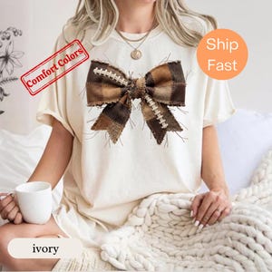 May include: An ivory short-sleeve t-shirt featuring a large, centred graphic of a brown and tan plaid bow. The bow design incorporates white football lace patterns across its fabric. The shirt has a crew neckline and a relaxed, loose fit.