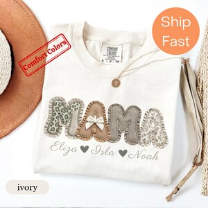 Puede incluir: Una camiseta de color marfil con la palabra MAMA en letras grandes, texturizadas y con volantes, que presentan varios patrones como estampado de leopardo, beige liso con un pequeño lazo blanco, espiga y flores. Debajo aparecen los nombres impresos.