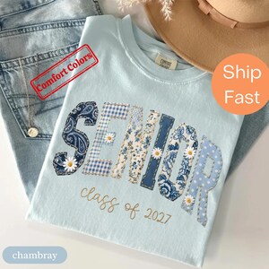 Puede incluir: Una camiseta azul claro con la palabra SENIOR en letras grandes de tela estampada con diseños de cachemira, cuadros vichy, flores y lunares azules, acentuadas con pequeñas margaritas blancas. Debajo, el texto class of 2027 está en letra dorada.