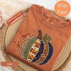 Puede incluir: Una camiseta de cuello redondo color óxido con un gran gráfico de calabaza compuesto por varios segmentos de tela estampada, incluyendo hojas de otoño, bellotas, un tejido verde, calabazas y un estampado de cielo nocturno azul oscuro.