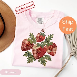 May include: A light pink t-shirt featuring a central graphic of three strawberries made from various red and white patterned fabric patches. Each strawberry is accented with a small, circular beaded flower and surrounded by green fabric leaves.