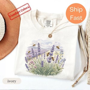 May include: An ivory t-shirt featuring a watercolor-style graphic of purple lavender fields, green foliage, and several honeybees in flight. The shirt has a crew neckline and a Comfort Colors brand label printed on the inside of the collar.