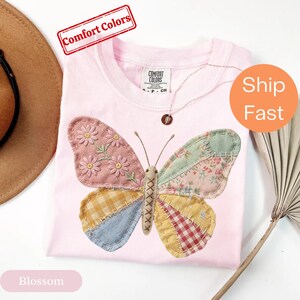 Puede incluir: Una camiseta rosa claro con una gran aplicación de mariposa hecha de varios parches de tela, incluyendo patrones florales, de cuadros vichy y lisos. La mariposa tiene detalles de margaritas bordadas en el ala superior izquierda y un cuerpo central.