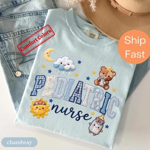 Può includere: Una t-shirt azzurra con la scritta PEDIATRIC nurse in lettere maiuscole a motivi blu e bianchi. Il design include un sole giallo, una luna crescente, una nuvola bianca, un orsacchiotto marrone e un biberon, decorati con piccole stelle.